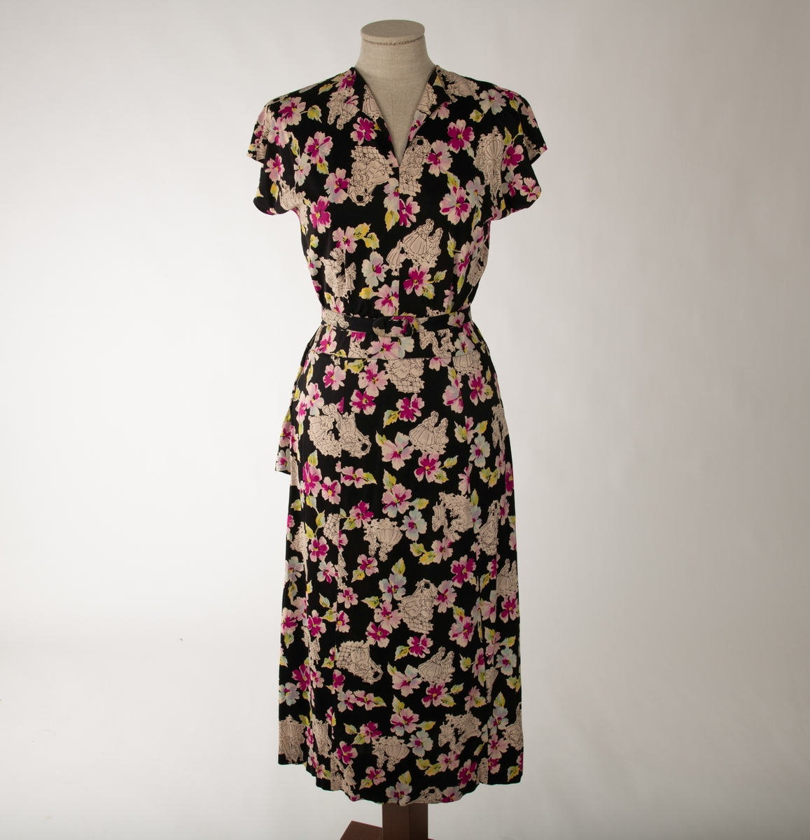 En svart kjole med et mangefarget motiv av blomster og en mann og en kvinne i ulike scener, begge kledd i 1700-talls overklasse-klær. Kjolen er ermeløs og krageløs. En glidelås under venstre arm går ned et lite stykke. Under denne er det et en splitt. Under halsen er det felt inn et svart gjennomsiktig stoff. Et hekte og en hempe i nakken skal lukke kjolen der, men hempe har røket

Det hører til et belte som er kledd i samme stoff.