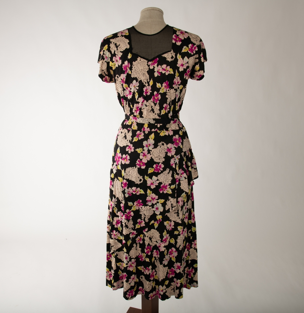 En svart kjole med et mangefarget motiv av blomster og en mann og en kvinne i ulike scener, begge kledd i 1700-talls overklasse-klær. Kjolen er ermeløs og krageløs. En glidelås under venstre arm går ned et lite stykke. Under denne er det et en splitt. Under halsen er det felt inn et svart gjennomsiktig stoff. Et hekte og en hempe i nakken skal lukke kjolen der, men hempe har røket

Det hører til et belte som er kledd i samme stoff.