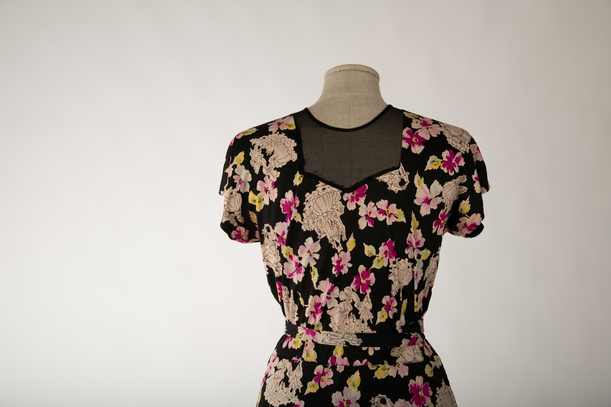 En svart kjole med et mangefarget motiv av blomster og en mann og en kvinne i ulike scener, begge kledd i 1700-talls overklasse-klær. Kjolen er ermeløs og krageløs. En glidelås under venstre arm går ned et lite stykke. Under denne er det et en splitt. Under halsen er det felt inn et svart gjennomsiktig stoff. Et hekte og en hempe i nakken skal lukke kjolen der, men hempe har røket

Det hører til et belte som er kledd i samme stoff.