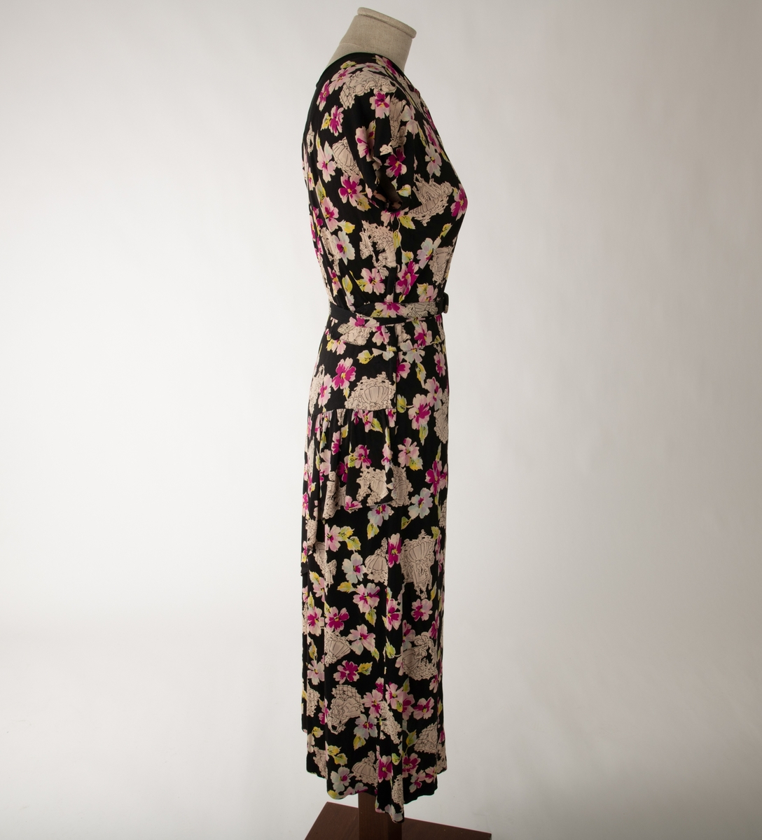 En svart kjole med et mangefarget motiv av blomster og en mann og en kvinne i ulike scener, begge kledd i 1700-talls overklasse-klær. Kjolen er ermeløs og krageløs. En glidelås under venstre arm går ned et lite stykke. Under denne er det et en splitt. Under halsen er det felt inn et svart gjennomsiktig stoff. Et hekte og en hempe i nakken skal lukke kjolen der, men hempe har røket

Det hører til et belte som er kledd i samme stoff.