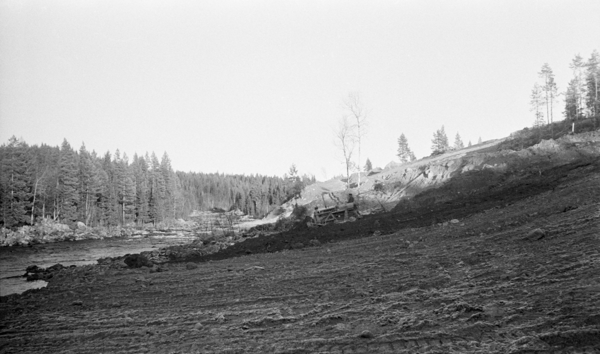 Opparbeiding av velteplass for fløtingstømmer ved elva Flisa i Åsnes i Solør i 1951. Fotografiet viser en bulldoser som jevner ut overflata på ei slaktskrånende helling nedover mot elveløpet. Tanken var nok at dette stedet skulle bli en tømmerterminal, en mottaksplass for tømmer som kunne hentes hit fra omliggende skoger med lastebiler via de mange skogsbilvegene som kom i etterkrigstida. På denne måten unngikk man den vanskelige fløtinga i de mange små sidevassdragene - bekker og åer med liten vannføring. Ved å slå tømmeret direkte på de større hovedvassdragene var man sikrere på å få det framfløtt uten at knapphet på vann forsinket eller stanset arbeidet.