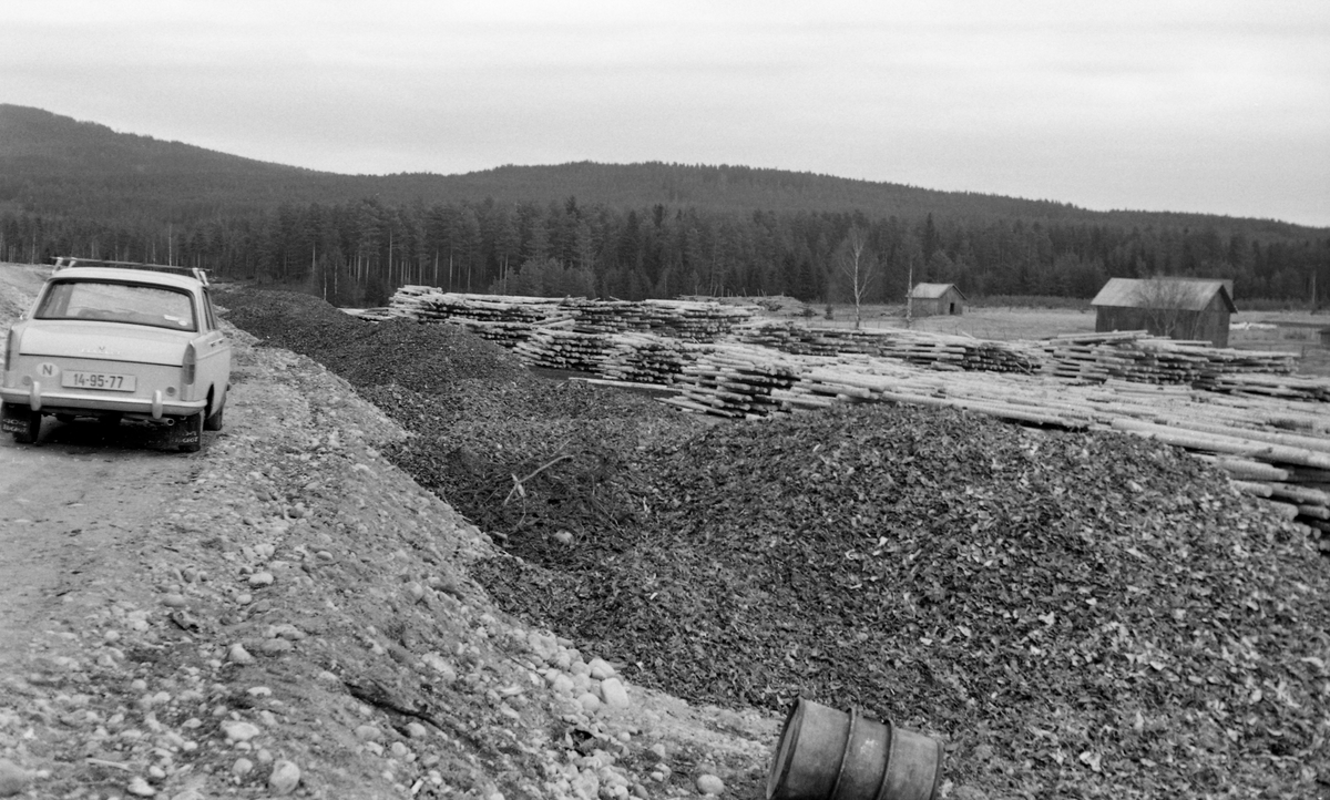 Fra Storvelta ved elva Flisa i Åsnes i Solør. Fotografiet ble tatt i 1964. Storvelta var en av mange velte- og terminalplasser Glomma fellesfløtingsforening fikk opparbeidet ved hovedårene i det vassdraget denne organisasjonen brukte som transportåre for tømmer. Disse oppsamlingsstedene for store tømmervolumer ble bygd etter at skogene i løpet av to-tre tiår etter 2. verdenskrig hadde fått et vidgreinet nett av skogsbilveger som gjorde det umulig å hente ut tømmer fra områder der virket tidligere hadde drevet ut via åer og bekker der fløtinga var arbeidskrevende og sårbar på grunn av begrenset tilgang på vann. Nå kunne tømmeret hentes med lastebiler fra velter ved de nevnte skogsbilvegene og kjøres til større velteplasser ved de større vassdragene. Store kvanta tømmer på slike steder gjorde det også interessant å investere i barkemaskiner. Vinterstid kunne manuell barking med øks eller barkespade legge beslag på 40-50 prosent av skogsarbeidernes arbeidstid. Her var det med andre ord både slit og arbeidstimer å spare. Storvelta ved Flisa fikk en ringbarkemaskin (jfr. SJF.1994-00825 og SJF.1994.00829). Dette fotografiet viser hvordan barken fra denne maskinen ble deponert langs bilvegen til velteplassen. Bak barkhaugene ser vi hauger av tømmer som var opplagt i strøvelter med sikte på at stokkene skulle få en forsiktig tørk, noe som gav dem bedre oppdrift i vannet når fløtingssesongen startet. I bakgrunnen skimter vi et par høyløer. Til venstre i bildet ser vi en personbil, en Peugeot.