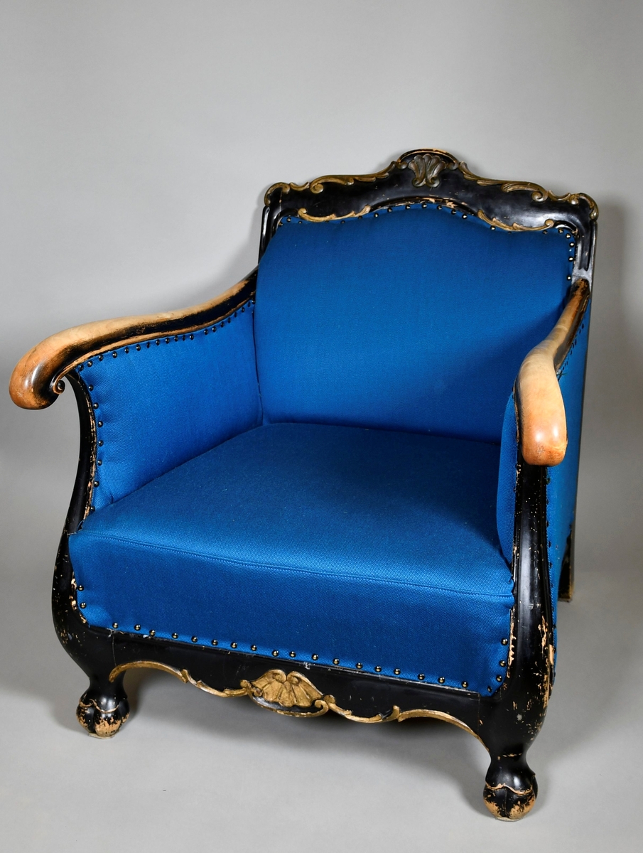 Barokkinspirert lenestol. Treverket har svart grunnfarge som er avslitt flere steder, spesielt på armlenene. Toppen av ryggen har symmetrisk skåret dekor av akantusranker, og et stående skjell i midten. Randen har bølgete form. Armlenene har kurvet form, og nederst på håndtakene er det volutter. Sprossen under setet har liggende skjell med to volutter på toppen. Treskåret dekor er gullfarget. Stolen har nytt, blått trekk.