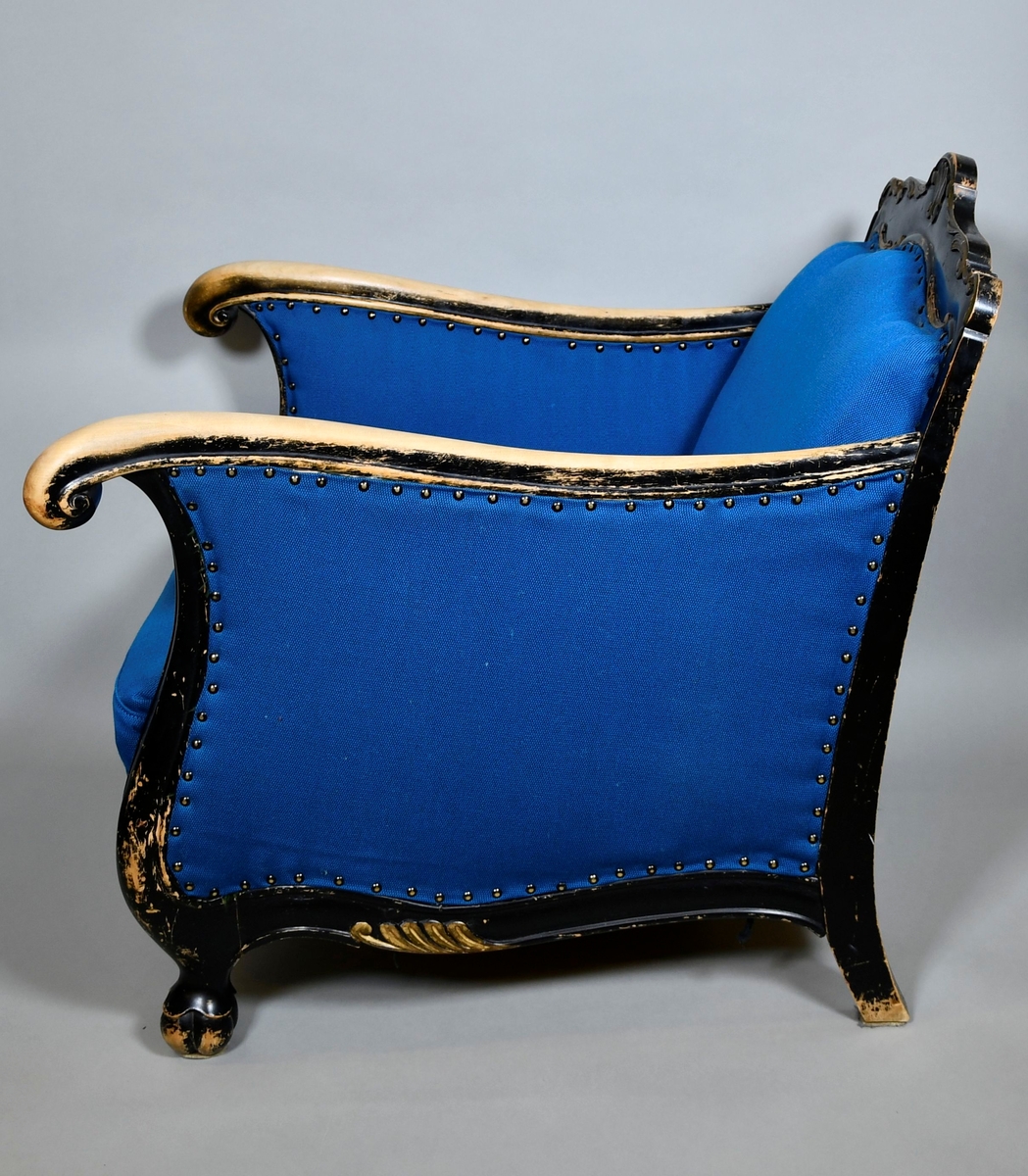 Barokkinspirert lenestol. Treverket har svart grunnfarge som er avslitt flere steder, spesielt på armlenene. Toppen av ryggen har symmetrisk skåret dekor av akantusranker, og et stående skjell i midten. Randen har bølgete form. Armlenene har kurvet form, og nederst på håndtakene er det volutter. Sprossen under setet har liggende skjell med to volutter på toppen. Treskåret dekor er gullfarget. Stolen har nytt, blått trekk.