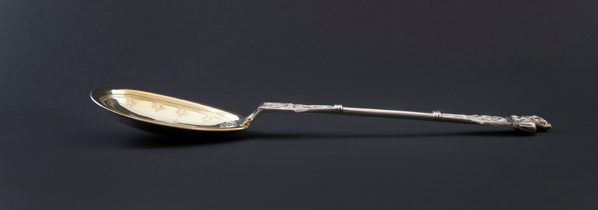 Kompottskje i sølv og dragestil. Det har ovalt blad og er innvendig gravert med vikingskip på forgylt bakgrunn. Baksiden er gravert med drageslyng i entrelac nær roten. Skaftroten har støpt dekor med drager i relieff. Likeledes er det utvidede toppstykket med liggende dyr som toppfigur. Dyrets hode er vendt en face med tungen ut av munnen. Skaftets midtparti har tynn, glatt utforming.