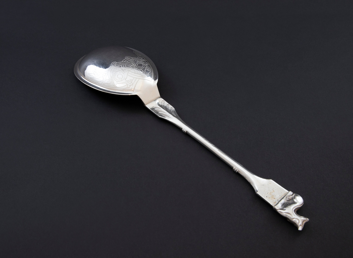 Kompottskje i sølv og dragestil. Det har ovalt blad og er innvendig gravert med vikingskip på forgylt bakgrunn. Baksiden er gravert med drageslyng i entrelac nær roten. Skaftroten har støpt dekor med drager i relieff. Likeledes er det utvidede toppstykket med liggende dyr som toppfigur. Dyrets hode er vendt en face med tungen ut av munnen. Skaftets midtparti har tynn, glatt utforming.