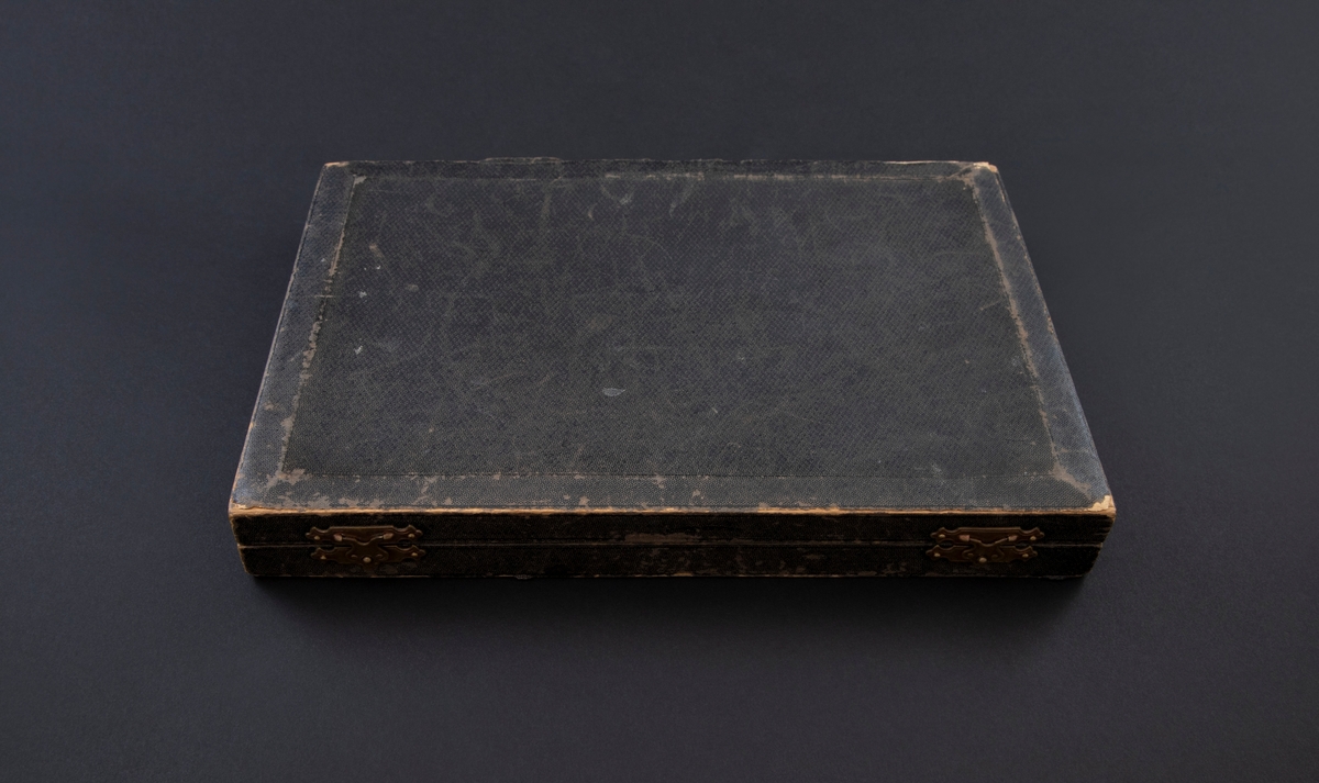 Etui av tre med hengslet lokk og to låser. Den er kledd med skinnlignende, svart papir. Innvendig er lokket kledd med lyseblått, silkelignende stoff og bunnen er fôret med kremgul polstring.