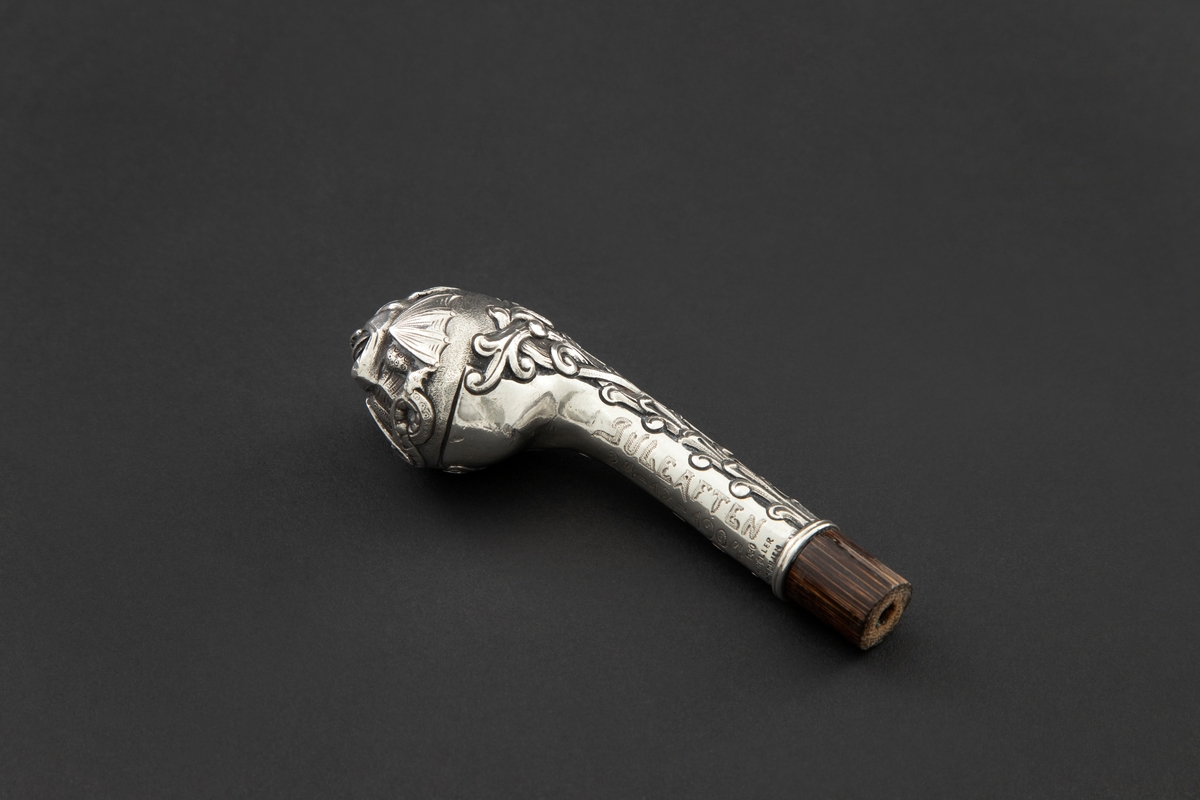 Skafthåndtak til paraply i sølv. Den er støpt med lett svunget hals som ender i en kuleformet avslutning. På kulen i relieff er en grotesk, et djevelhode, med forkropp, vinger og hale. Sidene på halsen er dekorert med romanske ranker. Holken er montert på en avkappet, hul treplugg.