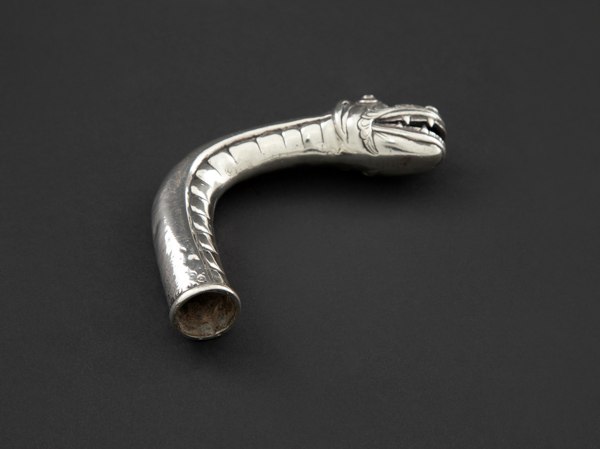 Skafthåndtak til paraply i sølv. Den er støpt, hul og formet som et dragehode med kraftig svungen hals. Halsen danner selve håndgrepet.