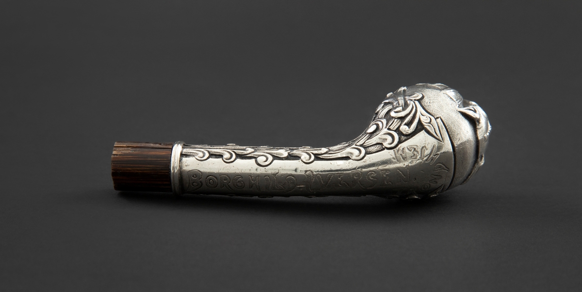 Skafthåndtak til paraply i sølv. Den er støpt med lett svunget hals som ender i en kuleformet avslutning. På kulen i relieff er en grotesk, et djevelhode, med forkropp, vinger og hale. Sidene på halsen er dekorert med romanske ranker. Holken er montert på en avkappet, hul treplugg.