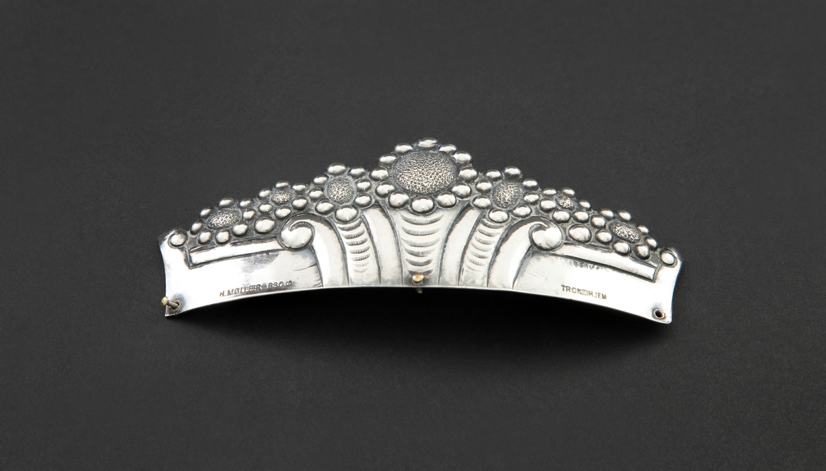 Øvre del av pyntekam i sølv, brukt som hårpynt. Den er drevet og siselert med flat, buet form. Dekoren består av syv blomster, eller rosetter, over et flatt felt med vertikale, organiske riller i midten. Langs nederste kant er tre hull for feste til kam, hvori to av tre nitter er i behold.