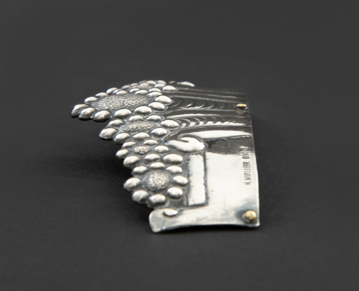 Øvre del av pyntekam i sølv, brukt som hårpynt. Den er drevet og siselert med flat, buet form. Dekoren består av syv blomster, eller rosetter, over et flatt felt med vertikale, organiske riller i midten. Langs nederste kant er tre hull for feste til kam, hvori to av tre nitter er i behold.