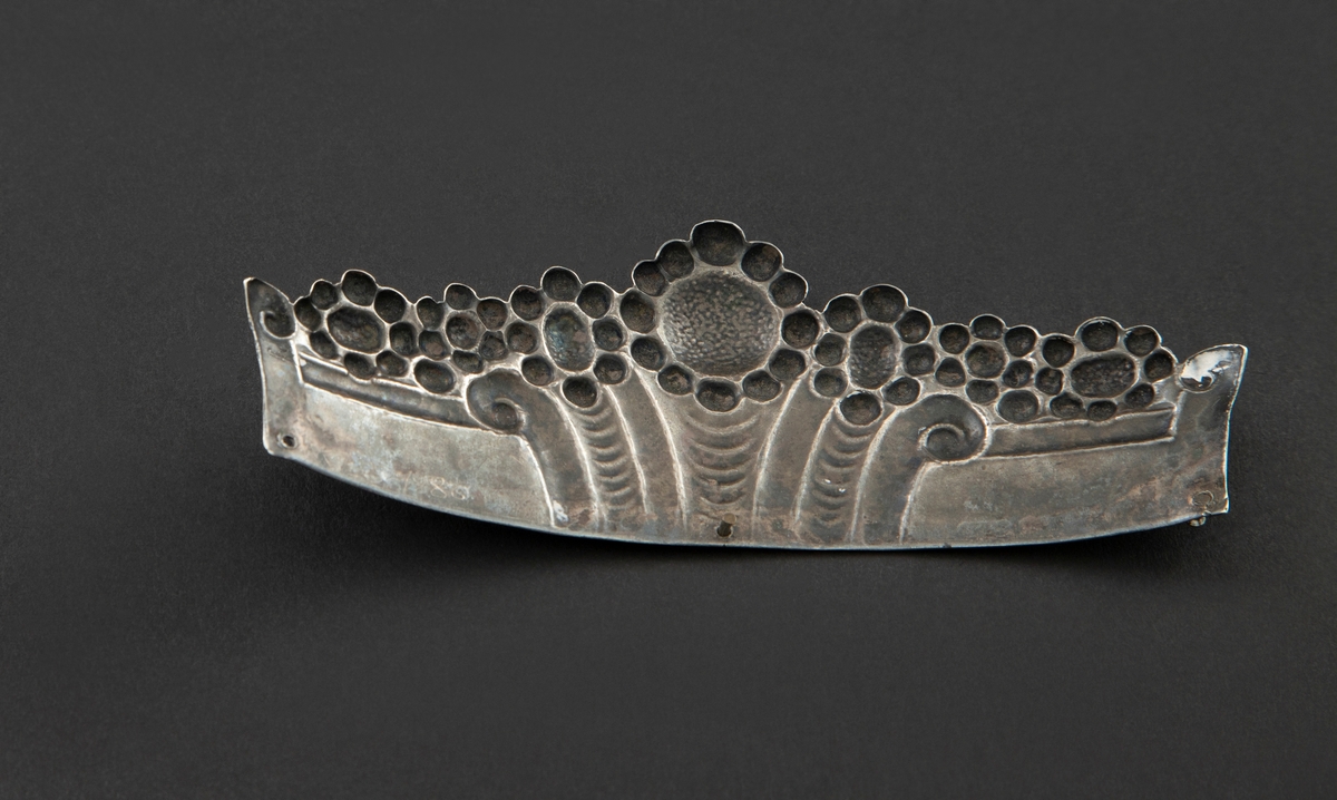 Øvre del av pyntekam i sølv, brukt som hårpynt. Den er drevet og siselert med flat, buet form. Dekoren består av syv blomster, eller rosetter, over et flatt felt med vertikale, organiske riller i midten. Langs nederste kant er tre hull for feste til kam, hvori to av tre nitter er i behold.