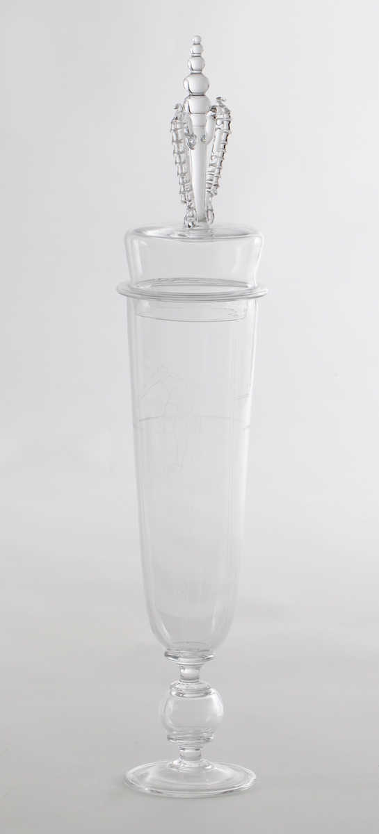 Pokal med lokk i klart glass, dekorert med gravering i form av stiliserte atleter. 

1) Høyreist sylinderformet korpus som hviler på en kuleformet stett, samt noe hevet sirkulær fotplate. På øvre del av korpus er det gravert en mannlig atlet som utfører friidrett, trolig stavsprang.

2) Tilnærmet sylinderformet lokk med høyreist kuleformet lokknapp, flankert av to mykt buede glasstaver med påklipp.