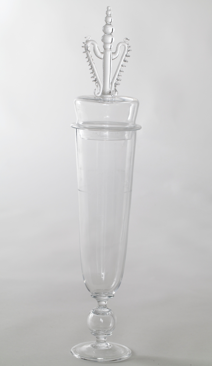 Pokal med lokk i klart glass, dekorert med gravering i form av stiliserte atleter. 

1) Høyreist sylinderformet korpus som hviler på en kuleformet stett, samt noe hevet sirkulær fotplate. På øvre del av korpus er det gravert en mannlig atlet som utfører friidrett, trolig stavsprang.

2) Tilnærmet sylinderformet lokk med høyreist kuleformet lokknapp, flankert av to mykt buede glasstaver med påklipp.