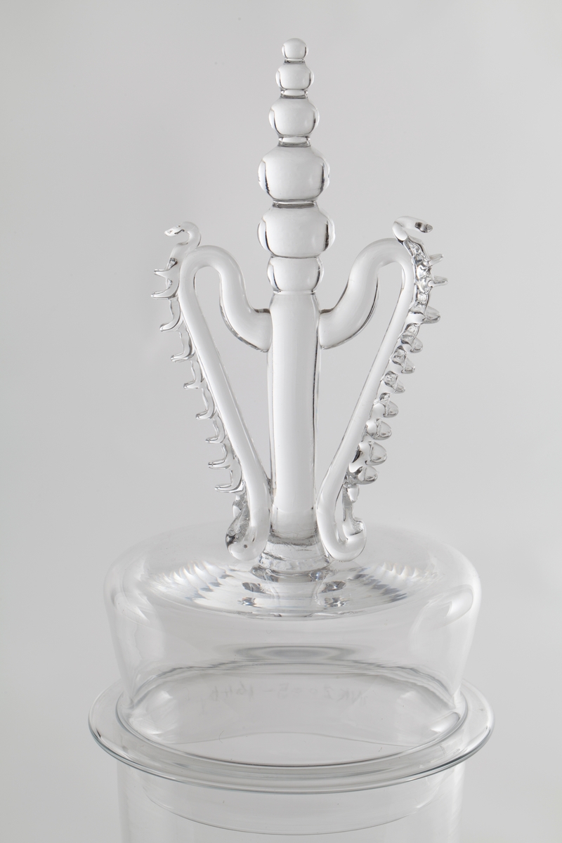 Tilnærmet sylinderformet lokk med høyreist kuleformet lokknapp, flankert av to mykt buede glasstaver med påklipp.