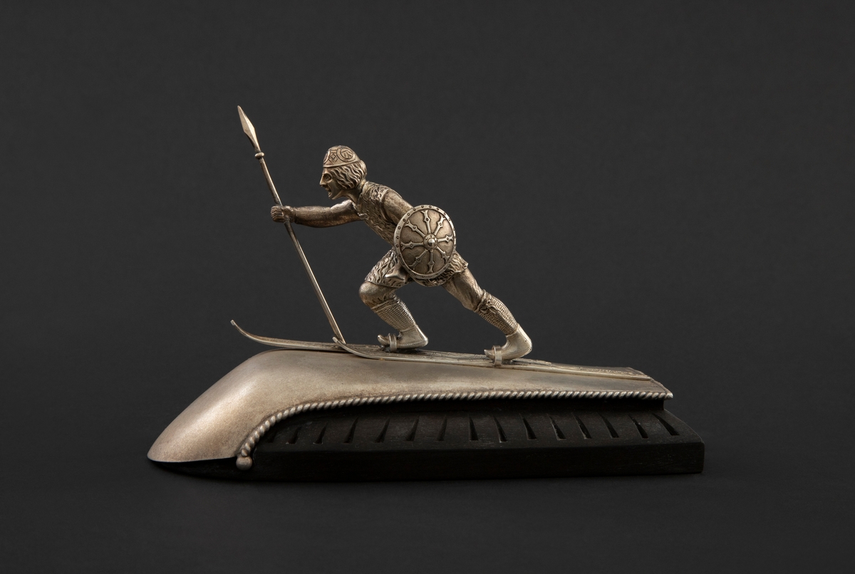 Statuett i støpt og siselert sølv på fotstykke av ibenholt. Statuetten forestiller en viking på ski med spyd i høyre og skjold i venstre hånd. Vikingen står på en krummet plate av matt - muligens syrebehandlet - sølv, som er skrudd fast til fotstykket.