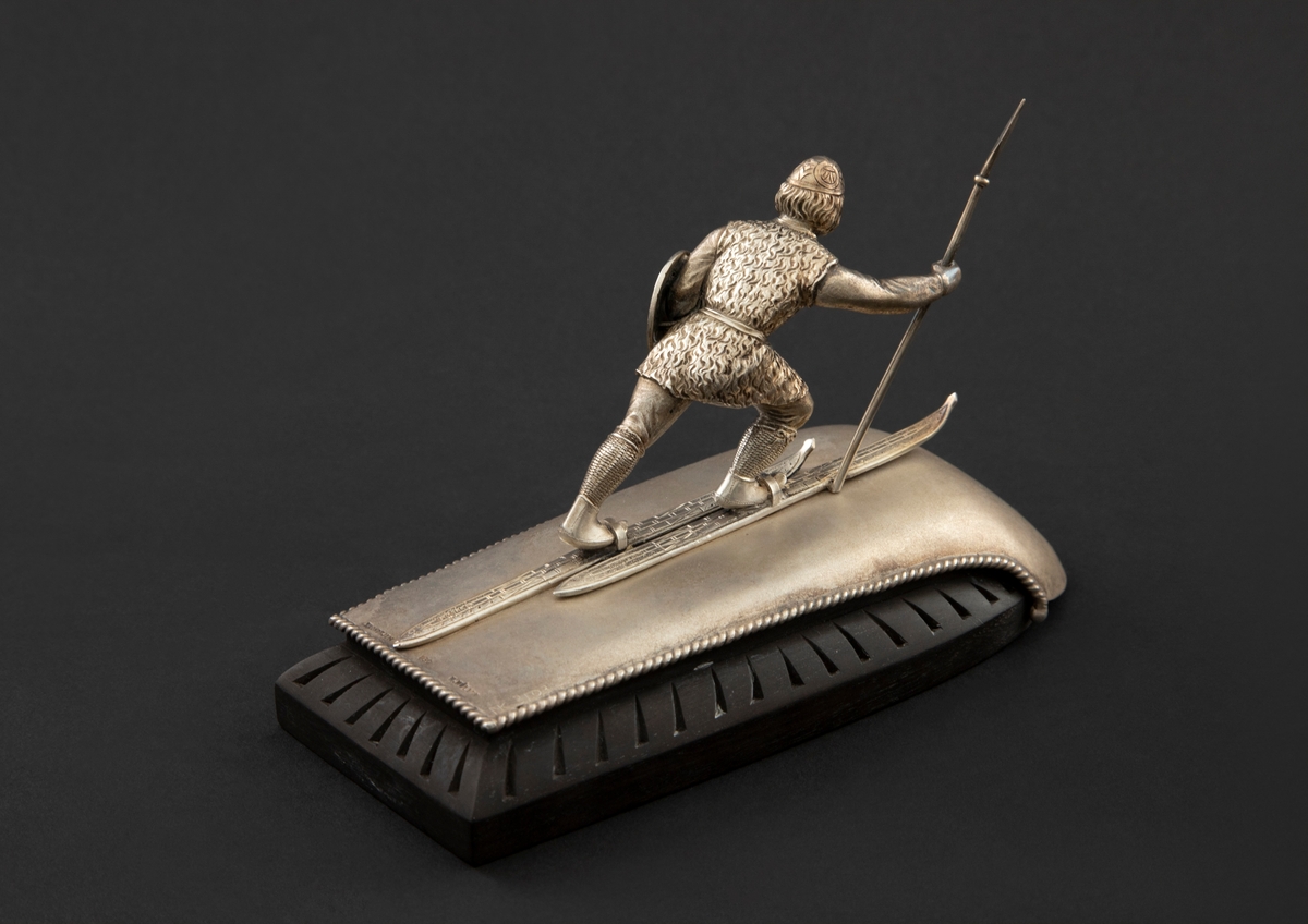 Statuett i støpt og siselert sølv på fotstykke av ibenholt. Statuetten forestiller en viking på ski med spyd i høyre og skjold i venstre hånd. Vikingen står på en krummet plate av matt - muligens syrebehandlet - sølv, som er skrudd fast til fotstykket.