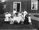 Fem kvinnor och två små flickor samlade runt kaffebordet på gräsmattan utanför Missionshuset i Uppgränna. Alla utom den äldre kvinnan till höger klädda i ljusa klänningar.
Den äldre kvinnan är Sofie Josefsson och två av kvinnorna är hennes döttar Agnes och Tyra, samt Agnes döttrar. Tyra står till vänster och Agnes står till höger.
