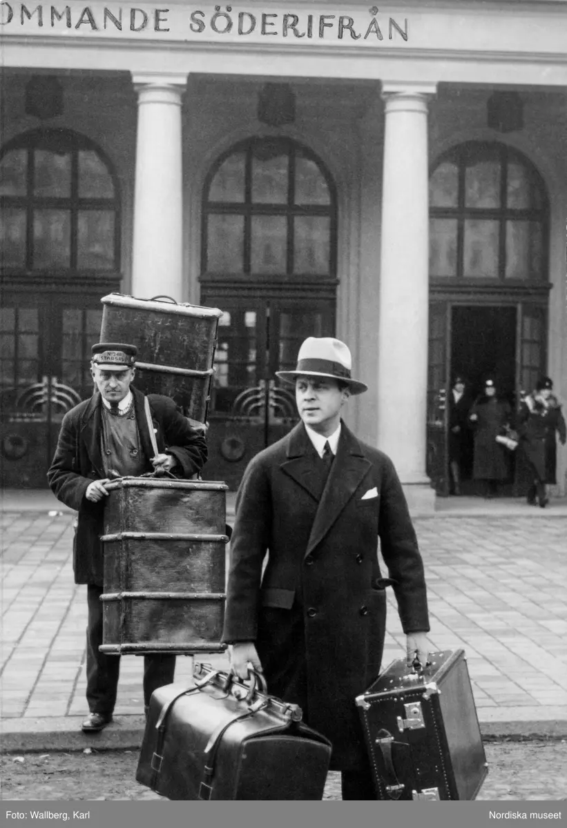 Två män framför Stockholms central, båda bär på bagage, resväskor och koffertar. Den ene mannen är ett stadsbud, den andre Kurt Jacobsson, chef för NK:s franska damskrädderi.