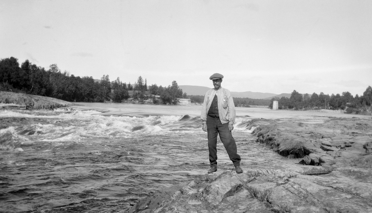 Edvard Lapstun (1875-1950) ved Eidsfossen i Glomma, i Våler i Hedmark i 1920. Lapstun sto på ei bergflate på østre elvebredd, kledd i varmelsbukser, mørk vest med en stripete busserull med åpen knepping på forstykket og med sixpencelue på hodet. Da dette fotografiet ble tatt var han 45 år gammel og bodde på det lille bruket Tangrønningen med kone og fire barn. I lokalsamfunnet var han nok best kjent i rollen som fløterhusbond. I bakgrunnen på dette bildet skimter vi karene til ei bru som var under bygging.