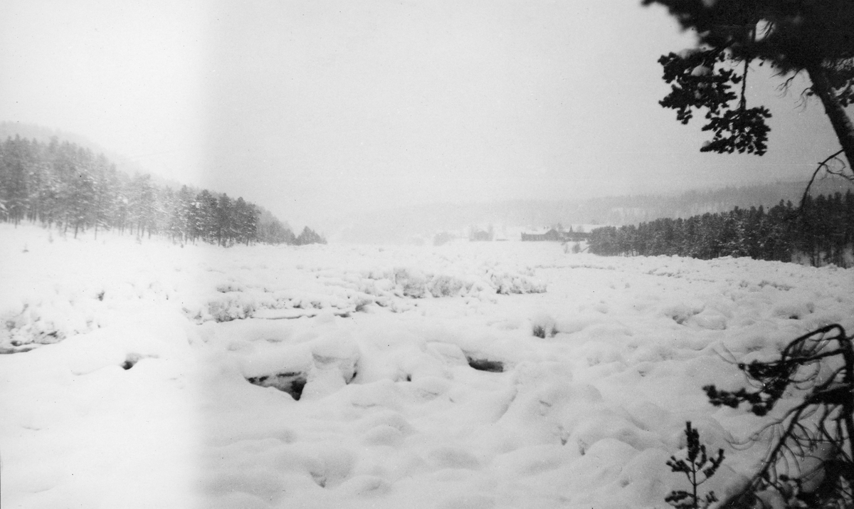 Tømmer under snø og is i Eidsfossen i Glomma, vinteren 1926. Eidsfossen ligger i Våler i Solør, Hedmark. I tåka til høye i bakgrunnen skimter vi antakelig tunet på en av Eid-gardene.