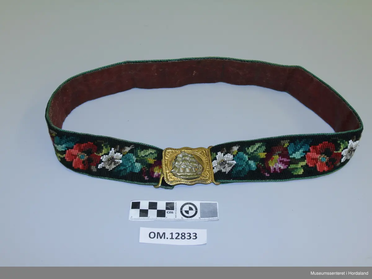 Beltet er brodert i kross-sting med ullgarn. Mønsteret er ulike blomster. Innimellom kvar blomster-rapport er det ein blomst i perlebroderi. Beltet er kanta med ei tvinna snor som er sydd på og fóra med eit raudt skinn. Spenna framme har eit skutemotiv som tilsei at det er eit mannsbelte.