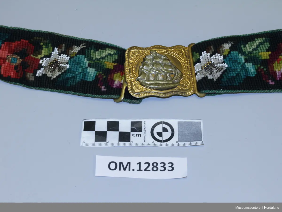Beltet er brodert i kross-sting med ullgarn. Mønsteret er ulike blomster. Innimellom kvar blomster-rapport er det ein blomst i perlebroderi. Beltet er kanta med ei tvinna snor som er sydd på og fóra med eit raudt skinn. Spenna framme har eit skutemotiv som tilsei at det er eit mannsbelte.