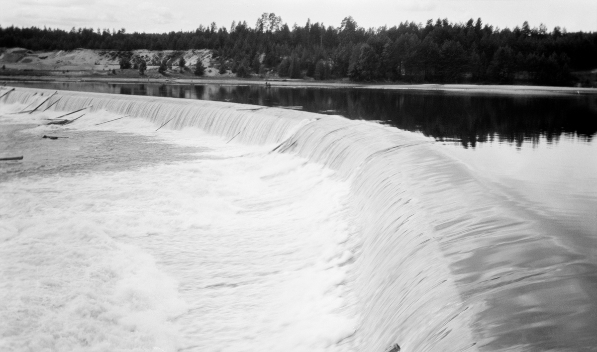 Dammen ved Skjefstadfossen i Elverum, fotografert fra den østre elvebredden med kameraet vendt mot den vestre sida av elva. Denne dammen ble bygd i 1909-1910 med sikte på at den skulle stuve opp vann til det nye kraftverket ved den vestre elvebredden i det ovenforliggende elveløpet. Dammen er om lag 300 meter lang, og den har en noe buet form. Damkrona ble støpt slik at den skrånet en aning nedover mot den østre enden. På den måten ønsket utbyggerne å trekke fløtingstømmeret mot den østre delen av elveløpet, for i vest lå kraftstasjonen og i midten av elva var det store steinører i sona nedenfor dammen, der tømmeret hadde lett for å sette seg fast. Da dette bildet ble tatt, var det enkelte stokker som hadde blitt stående i fallsona, særlig mot det nevnte grunne partiet i den midtre delen av elveløpet.