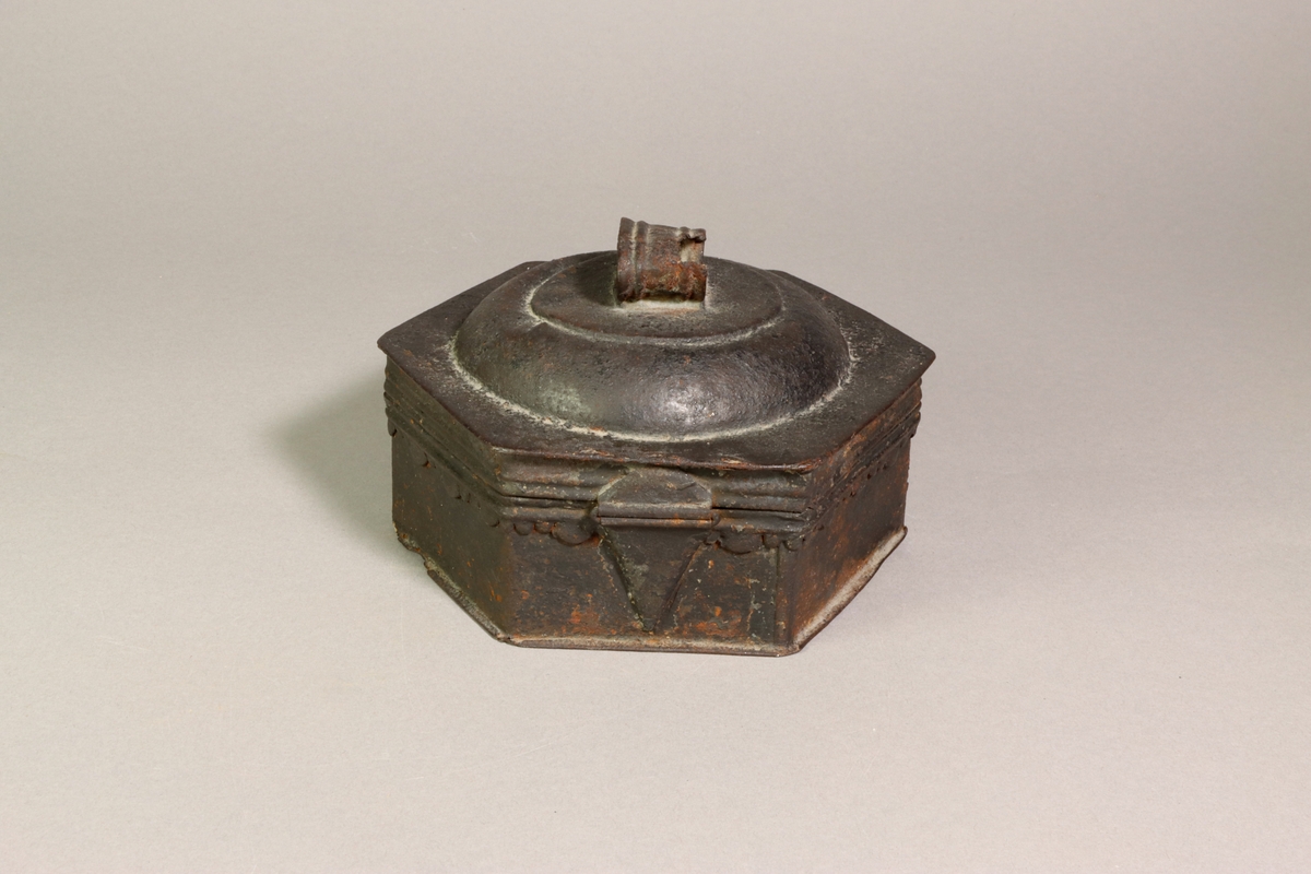 Skrin av metall med sexkantig form. Välvt lock krönt av en ögla. Locket är baktill fäst med ett gångjärn. Skrinets sidor är dekorerad med en lambrekäng. Fram på skrinet finns en hänkel för låsning. I skrinet finns sex stycken triangelformade fack som omger en rund hållare för ett rivjärn.