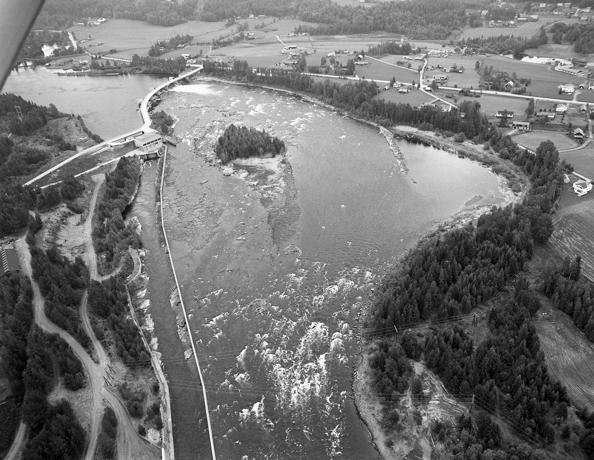 Flyfotografi, tatt over Glomma, fra et punkt like sør for Skjefstadfossen i Heradsbygda i Elverum kommune. Dette bildet skal være tatt på forsommeren i 1985, som ble den siste fløtingssesongen i dette vassdraget. Øverst i det venstre hjørnet av bildeflata ser vi damanlegget, som stuvet opp vannet i den ovenforliggende delen av elveløpet i dermed sikret god og stabil tilførsel av driftsvann til kraftstasjonene ved vestre elvebredd. Den første av disse, som ble bygd i 1909-10, er ikke synlig på fotografiet. I begynnelsen av 1960-åra ble det konstatert at både dammen og kanalmuren begynte å bli svært forvitret. Vassdragsdirektoratet forutsatte fornyelse. Derfor ble det bygd en ny såkalt platedam noen få meter ovenfor den opprinnelige i 1965-66. Arbeidet ble utført av entreprenørselskapet A/S Høyer Ellefsen. I 1969 engasjerte Elverum elektrisitetsverk ingeniørfirmaet Elliot Strømme og arkitekt Egil Sorteberg for å få hjelp til å planlegge en helt ny kraftstasjon ved Skjefstadfossen. Elliot Strømme foreslo å øke fallhøyden fra 10,5 til 12 meter med sikte på å installere et kaplanaggregat en som forutsatte en vanngjennomstrømming på 180 kubikkmeter i sekundet. Dette skulle i så fall kunne gi en produksjon på120 millioner kWh - mer en tre ganger så stor kapasitet som den gamle kraftstasjonen hadde. Prosjektet fikk konsesjon i kraft av kongelig resolusjon av 23. juli 1971. Byggearbeidene ble utført av entreprenørselskapet Ingeniør F. Selmer AS i 1972-73. Skjefstadfoss kraftverk II leverte strøm alt fra november 1972. Den gamle kraftstasjonen var fortsatt i drift i perioder med rikelig vannføring. Etter at den nye kraftstasjonen og dammen var ferdig ønsket Elverum kommune å bygge vegbru over Glomma på dammen, for dermed å etablere en kjørbar forbindelse mellom Vestsivegen og Solørvegen. Dette prosjektet ble realisert som en videreføring av Elverum elektrisitetsverks utbyggingsarbeid. Fløtingstømmeret fra den ovenforliggende delen av Glommavassdraget ble - ved hjelp av lenser - ledet mot ei av de seks klapplukene i dammen, med sikte på at det skulle gå i den østre delen av elveløpet, der det var djupere og mindre oppstikkende berg og steinblokker enn på østsida av Tjuvholmen. Bildet viser at det var bygd en skjerm som skulle forebygge at myew av fløtingsvirket drev inn i vika på den østre elvebredden, 5-600 meter nedenfor dammen. Nærmere den vestre elvebredden ble det støpt en lang betongmur som skulle skjerme avløpskanalen fra den nye kraftstasjonen fra det øvrige elvevannet. I bakgrunnen, øverst på bildet, ser vi noe av bebyggelsen på den flate elvelsletta lngst nord i Heradsbygda.