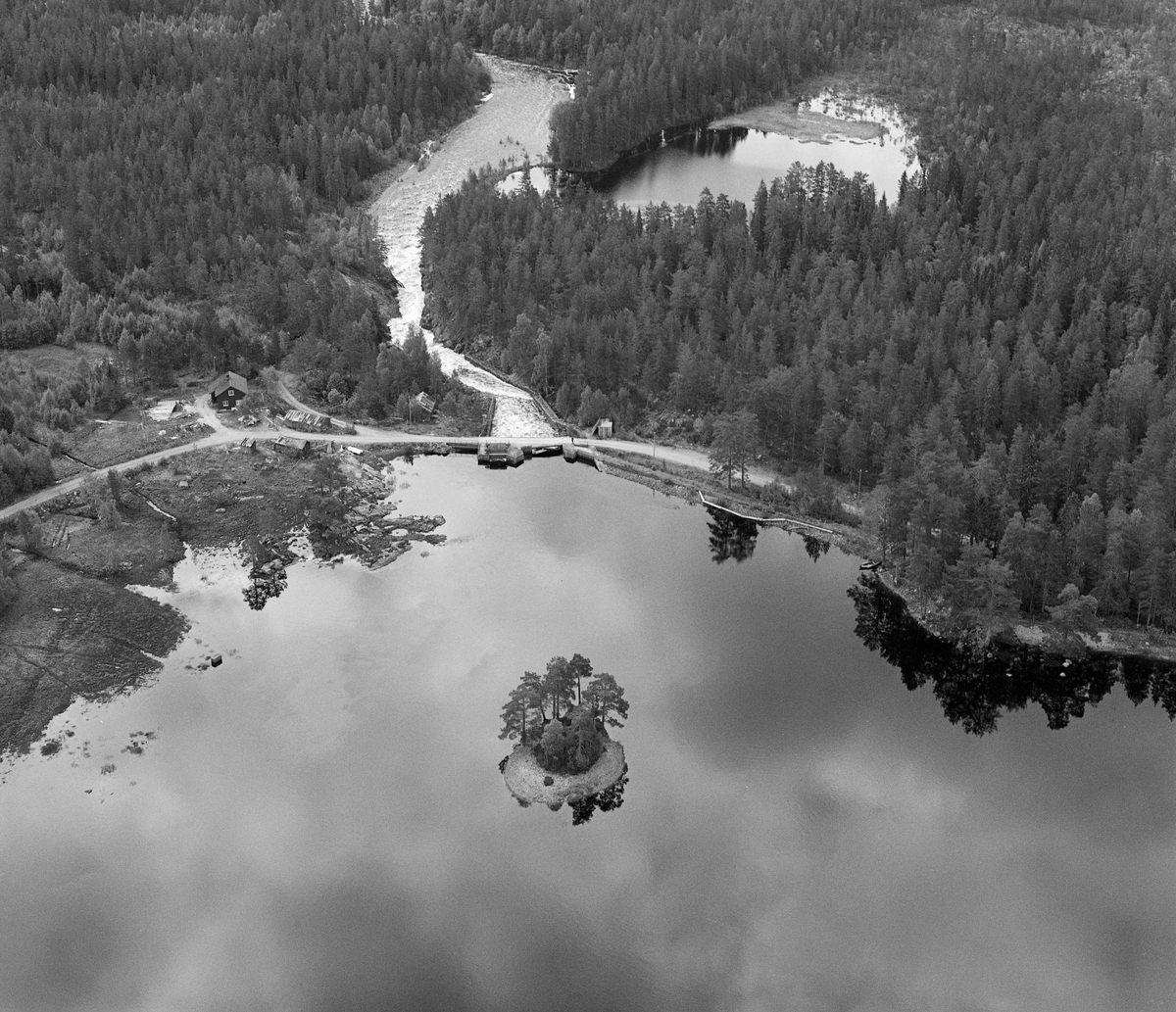 Flyfoto fra Haldammen, ved Halåas utløp i sørenden av Halsjøen. Denne lokaliteten ligger lengst øst i Våler kommune i Hedmark, snaut 300 meter over havet, og den nordøstre enden av den drøyt 4 kvadratkilometer store sjøen befinner seg på svensk side av riksgrensa. I tømmerfløtingssammenheng fungerte Halsjøen både som oppsamlingssted for tømmer som i løpet av vintersesongene ble drevet ut av skogene i dette området og som vannmagasin for fløtinga i det nedenforliggende Flisavassdraget. Dammen vi skimter sentralt i dette bildet ble bygd i 1920-21. Det er en solid konstruksjon, utfort i hoggen naturstein. Med den ble det mulig å heve vannspeilet cirka 2 meter og dermed øke sjøens kapasitet som vannmagasin. Sjølve dammen hadde tre løp: Mot det østre landet (til venstre på dette bildet) var det et tappeløp, tømmerløpet befant seg i midten og helt til høyre var det et flomløp. Når det ble fløtet tømmer fra Halsjøen ble det lagt ei traktformet lense mot tømmerløpet. Umiddelbart nedenfor dammen var det et berglendt og ganske bratt parti, der vannet og fløtingstømmeret ble ledet mellom to kraftige «skådammer» - ledeskjermer - som var murt av hoggen naturstein som var delvis betongforblendet. På begge sider av dammen var det mannskapskoier for fløterne - navnene «Fyllerud» og «Spritfritt» indikerte at fløterne grupperte seg i to leirer i en periode da tilgjengelighet til alkohol var et omstridt samfunnsspørsmål. Da dette fotografiet ble tatt bodde damvokteren Auden Haldammen (1917-1996) fortsatt i våningshuset på østsida av dammen (til venstre på bildet). Tjernet like nedenfor dammen kalles Kverntjernet. Grenda Gravberget ligger 4-5 kilometer nordvest for Haldammen. Jorda der er skrinn, og med ett unntak har brukene forholdsvis lite dyrket mark. I tider da det gjaldt å være mest mulig sjølberget med mest mulig var det viktig å få malt kornavlingene uten å måtte reise for langt. Ved Halåa var det vannkraft også til dette formålet.