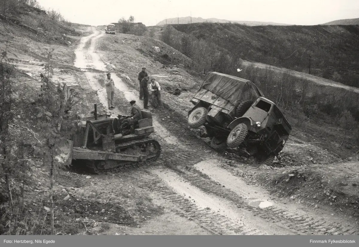 R.v. 50. 5 km. vest for Kunes, Laksefjord, 1948. Leirbakken. Det var gått et leirras tidligere og veglinja ble flyttet lengere ut fra bakken. Masseflyttingen ble foretatt med Bulldoser. En militærbil har kjørt ut fra veien. Vegskjæringen er stemplet av med planker. Veglinja forbi rasstedet. Skråningen gled ut og bilen seg ned i skråningen. Ingen ble skadet. Militærbilen prøver å trekke Bulldoseren tilbake på veien. Kåre Foslund er maskinfører. En ser vegen forbi rasstedet. Vegvesenets varevogn på toppen. Den nye vegfyllingen ser en til høyre. Lastebilen er en Fordson WOT. 