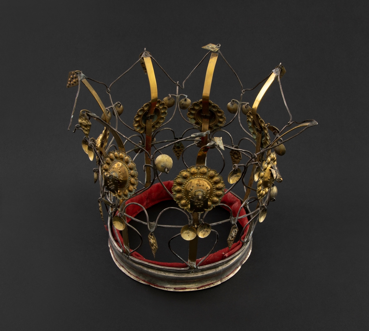 Brudekrone i sølv og messing. Den har vid, profilert bunnring, skjelett av sølvtråd og sju høye, vertikale standere av messing. Til hver av disse er festet en stor rosett av drevet messing. Over det hele er skjelettet dekorert med delvis forgylte anheng i form av blad, skåler og kors, drevet med bukler og masker. Bunnringen er innvendig fôret med rødt stoff.