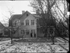 Fasadarbete pågår på större hus utmed Brahegatan i Gränna. Fotografi från gårdssidan, det ligger lite snö på gräsmattan. Två flickor står vinterklädda vid huset. Troligen barn till ägaren Björk.