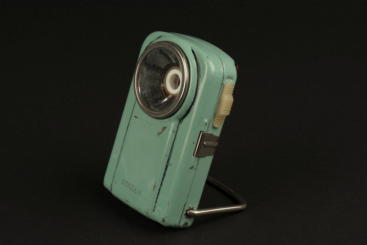 Rektangulär ficklampa med hölje av grönlackerad metall. 
På framsidan ett runt glas framför den nu saknade glödlampan. På baksidan en rektangulär röd reflex. På baksidan finns ett utfällbart metallstöd, som gör att lampan kan ställas.
Inuti finns hållare för ett batteri (saknas). Någon har skrivit både namn och ritat ett ansikte i tusch på insidan. Och på framsidan har svart tusch ritats i skarven mot den upphöjda mittdelen.