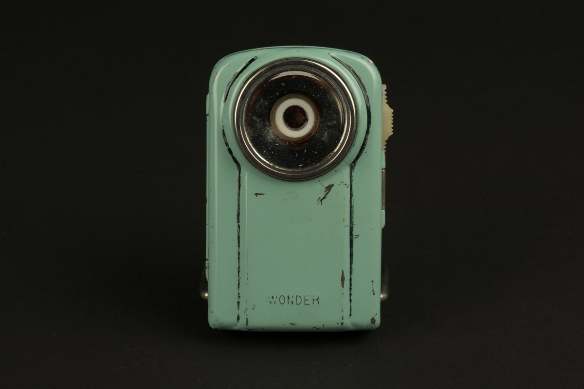 Rektangulär ficklampa med hölje av grönlackerad metall. 
På framsidan ett runt glas framför den nu saknade glödlampan. På baksidan en rektangulär röd reflex. På baksidan finns ett utfällbart metallstöd, som gör att lampan kan ställas.
Inuti finns hållare för ett batteri (saknas). Någon har skrivit både namn och ritat ett ansikte i tusch på insidan. Och på framsidan har svart tusch ritats i skarven mot den upphöjda mittdelen.