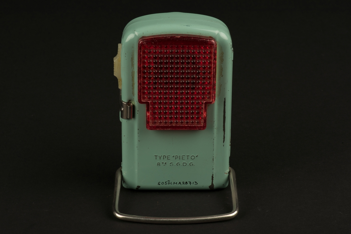 Rektangulär ficklampa med hölje av grönlackerad metall. 
På framsidan ett runt glas framför den nu saknade glödlampan. På baksidan en rektangulär röd reflex. På baksidan finns ett utfällbart metallstöd, som gör att lampan kan ställas.
Inuti finns hållare för ett batteri (saknas). Någon har skrivit både namn och ritat ett ansikte i tusch på insidan. Och på framsidan har svart tusch ritats i skarven mot den upphöjda mittdelen.