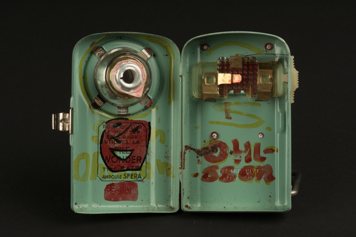 Rektangulär ficklampa med hölje av grönlackerad metall. 
På framsidan ett runt glas framför den nu saknade glödlampan. På baksidan en rektangulär röd reflex. På baksidan finns ett utfällbart metallstöd, som gör att lampan kan ställas.
Inuti finns hållare för ett batteri (saknas). Någon har skrivit både namn och ritat ett ansikte i tusch på insidan. Och på framsidan har svart tusch ritats i skarven mot den upphöjda mittdelen.