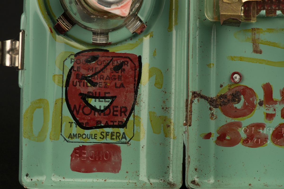 Rektangulär ficklampa med hölje av grönlackerad metall. 
På framsidan ett runt glas framför den nu saknade glödlampan. På baksidan en rektangulär röd reflex. På baksidan finns ett utfällbart metallstöd, som gör att lampan kan ställas.
Inuti finns hållare för ett batteri (saknas). Någon har skrivit både namn och ritat ett ansikte i tusch på insidan. Och på framsidan har svart tusch ritats i skarven mot den upphöjda mittdelen.