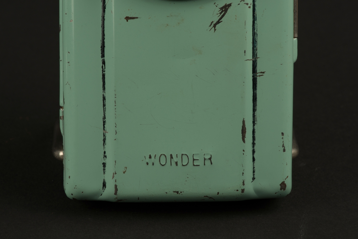 Rektangulär ficklampa med hölje av grönlackerad metall. 
På framsidan ett runt glas framför den nu saknade glödlampan. På baksidan en rektangulär röd reflex. På baksidan finns ett utfällbart metallstöd, som gör att lampan kan ställas.
Inuti finns hållare för ett batteri (saknas). Någon har skrivit både namn och ritat ett ansikte i tusch på insidan. Och på framsidan har svart tusch ritats i skarven mot den upphöjda mittdelen.