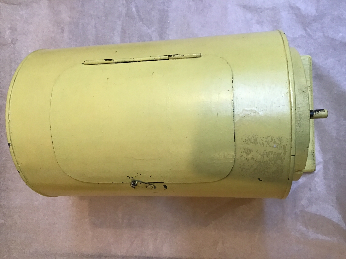 Lukket sylinder av gulmalt blikk som hviler i et stativ og kan sveives rundt. På siden er det en luke med enkel hendellukning. Ved mottak låg det en mengde blå papirlodd inni sylinderen.