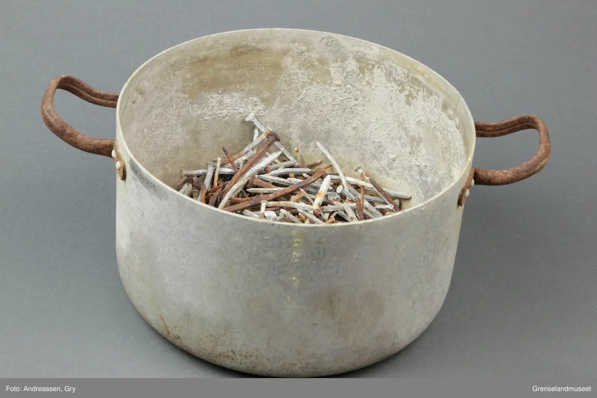 Rund aluminiumskasserolle med to små håndtak i annet metall. Håndtakene er rustet. 
I kjelen ligger det en stor mengde brukt spiker av varierende størrelse og type. Det er også en krok med øyekrok, en bolt, en strikkepinne og to smidde spiker, en lite øyekrok samt del av sluk til fiske. 