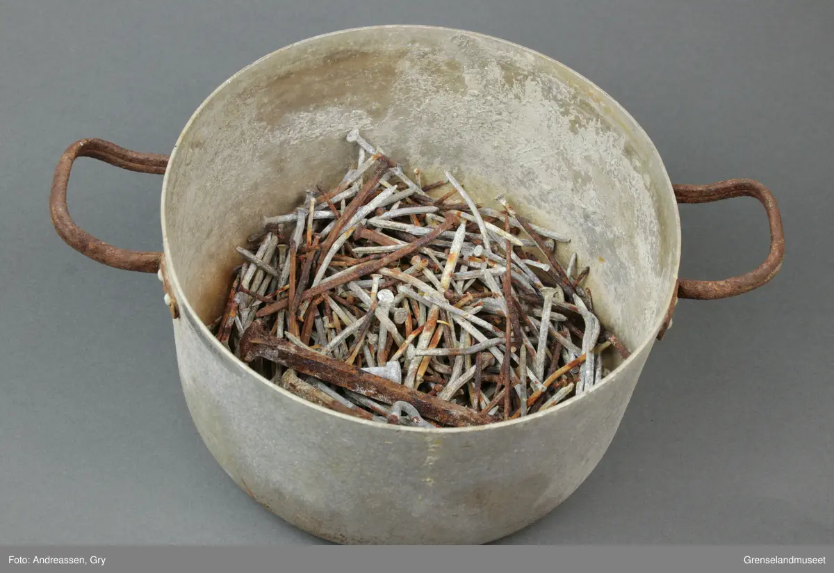 Rund aluminiumskasserolle med to små håndtak i annet metall. Håndtakene er rustet. 
I kjelen ligger det en stor mengde brukt spiker av varierende størrelse og type. Det er også en krok med øyekrok, en bolt, en strikkepinne og to smidde spiker, en lite øyekrok samt del av sluk til fiske. 