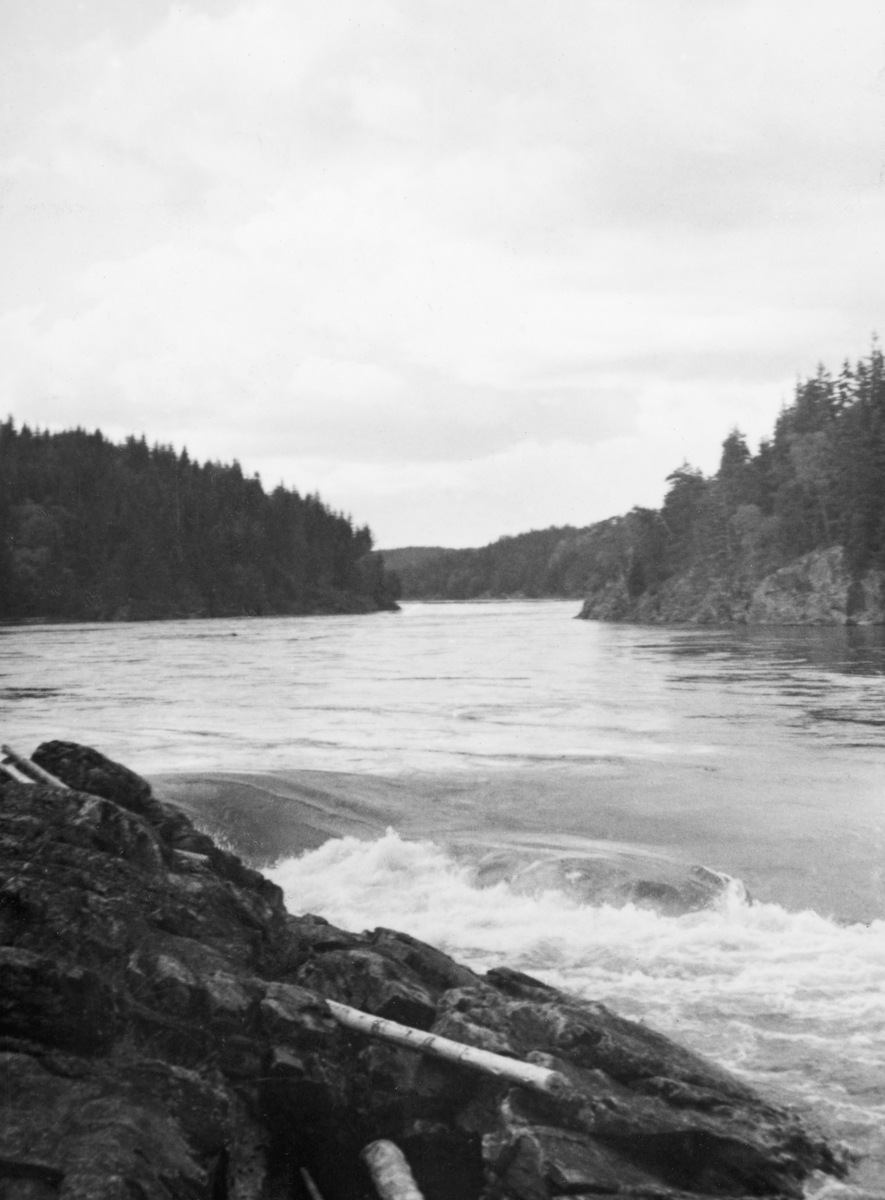 Smalt elveløp med fossestryk i Nedre Glomma. Bildet skal være tatt et sted mellom Kykkelsrud og Vamma i Østfold. Fotografen har stått på en bergnabb, ved sida av et sted der elvevannet faller over en terskel slik at det dannes en liten foss (til høyre i forgrunnen). Oppover langs elveløpet vokste det skog helt ned til vannkanten. Fotografiet kan være fra det elvestykket som kanaldirektør Gunnar Sætren tidlig på 1900-tallet beskrev slik: «Derpaa [nedenfor Vervenfossen] indsnævres elveleiet paany, og noget længere nede haves først Erterrisstryket og derpaa en række fosse med tilsammen 16 m. fald. De kaldes nu i almindelighed Vrangfos, men kaldtes tidligere Alvumfos, Vrangenfos, Helifos og Skjoldenfos efter de tilstødende Gaarde.»