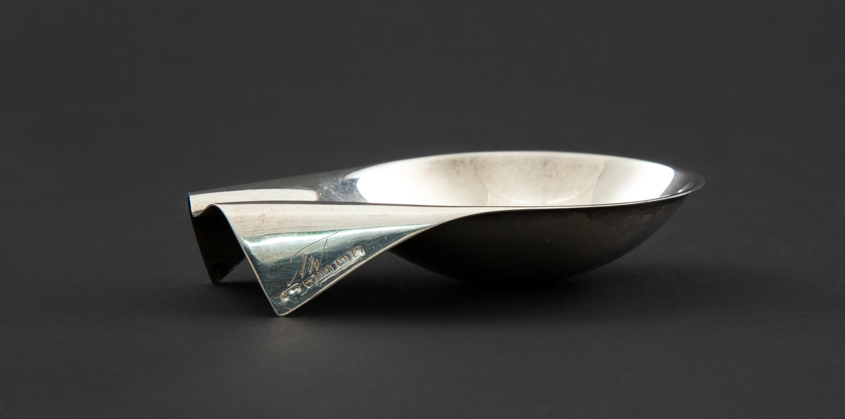 Pipestativ i sølv. Dråpeformet skål som smalner sammen til en forlenget endeparti med to nedbrettede kantfolder med monogram.