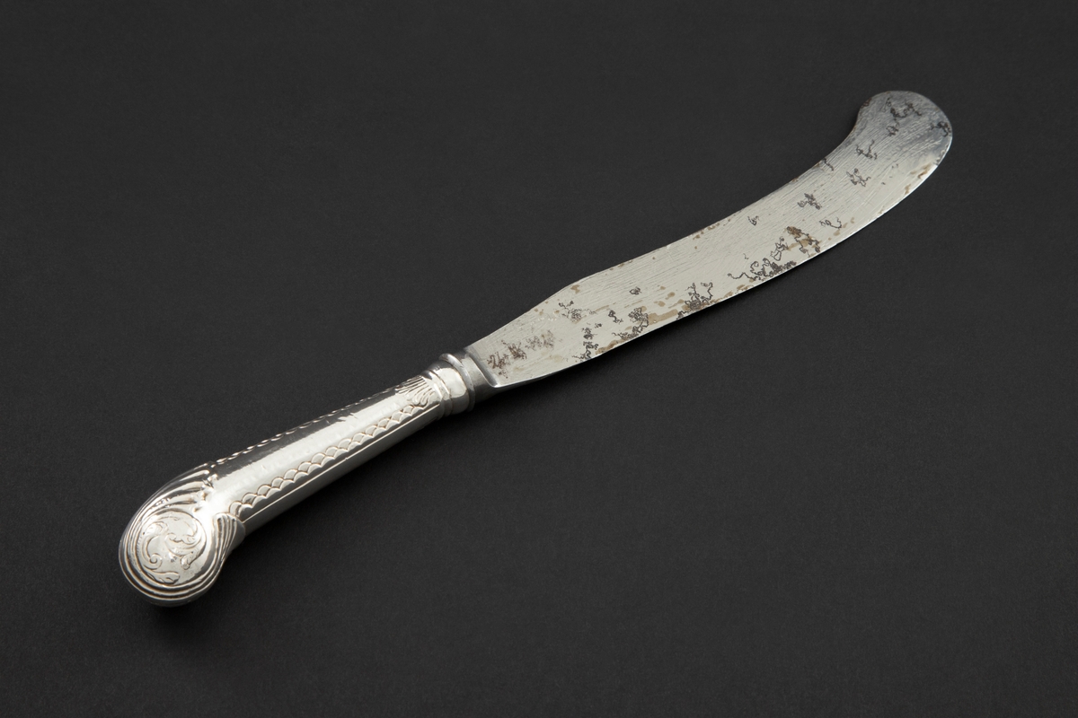 Kniv med sølvbelagt skaft og knivblad av stål. Knivbladet er flatt, buet med rund avslutning. Skaftet er rundt og dekorert med palmett- og bølgeornamentikk. Enden av skaftet er formet som et skjell med planteornamentikk.

