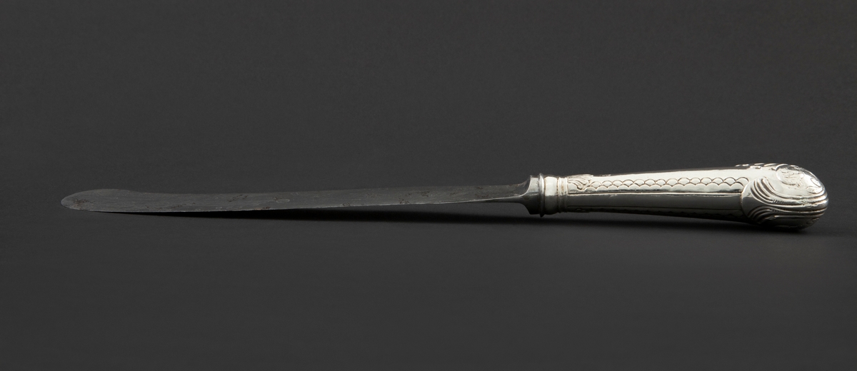 Kniv med sølvbelagt skaft og knivblad av stål. Knivbladet er flatt, buet med rund avslutning. Skaftet er rundt og dekorert med palmett- og bølgeornamentikk. Enden av skaftet er formet som et skjell med planteornamentikk.
