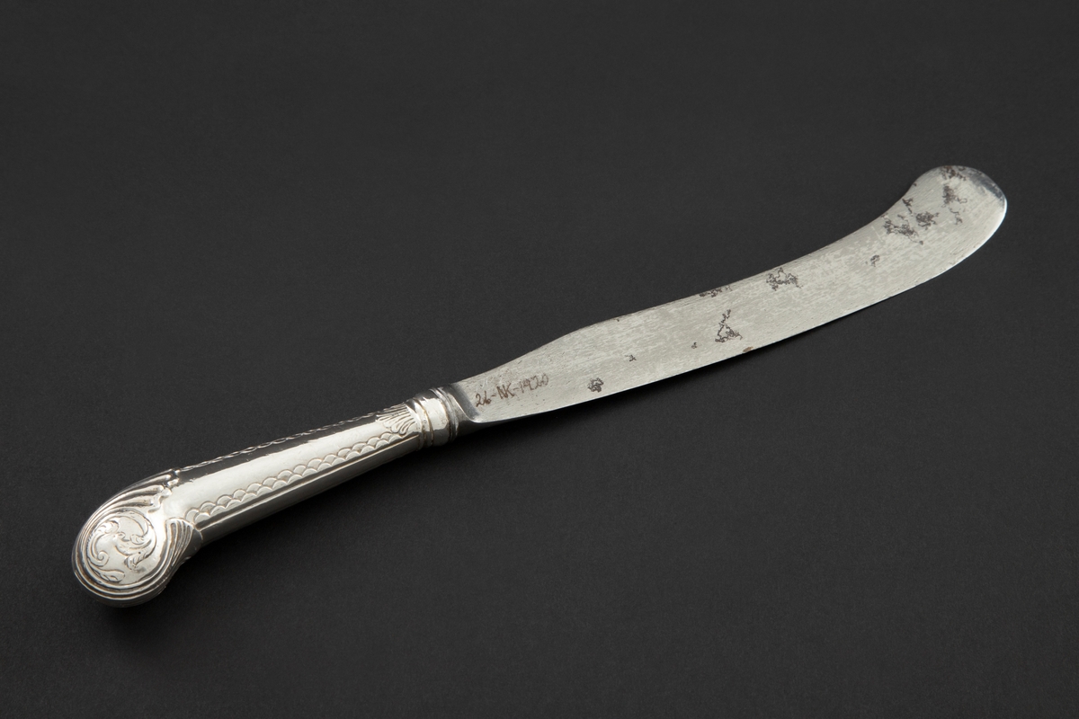 Kniv med sølvbelagt skaft og knivblad av stål. Knivbladet er flatt, buet med rund avslutning. Skaftet er rundt og dekorert med palmett- og bølgeornamentikk. Enden av skaftet er formet som et skjell med planteornamentikk.
