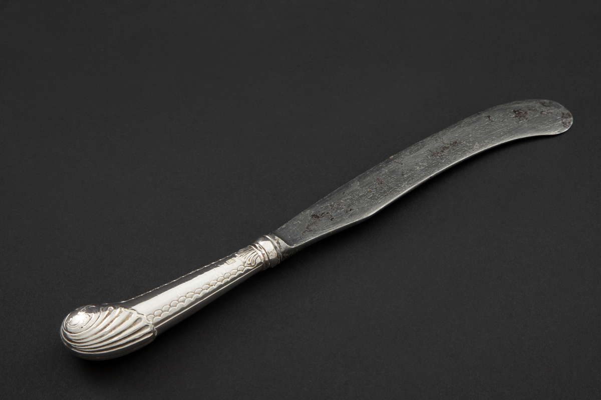 Kniv med sølvbelagt skaft og knivblad av stål. Knivbladet er flatt, buet med rund avslutning. Skaftet er rundt og dekorert med palmett- og bølgeornamentikk. Enden av skaftet er formet som et skjell med planteornamentikk.
